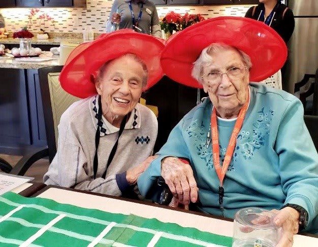 Two elderly women wearing red hats, sitting together and smiling, possibly at a celebration or gathering.