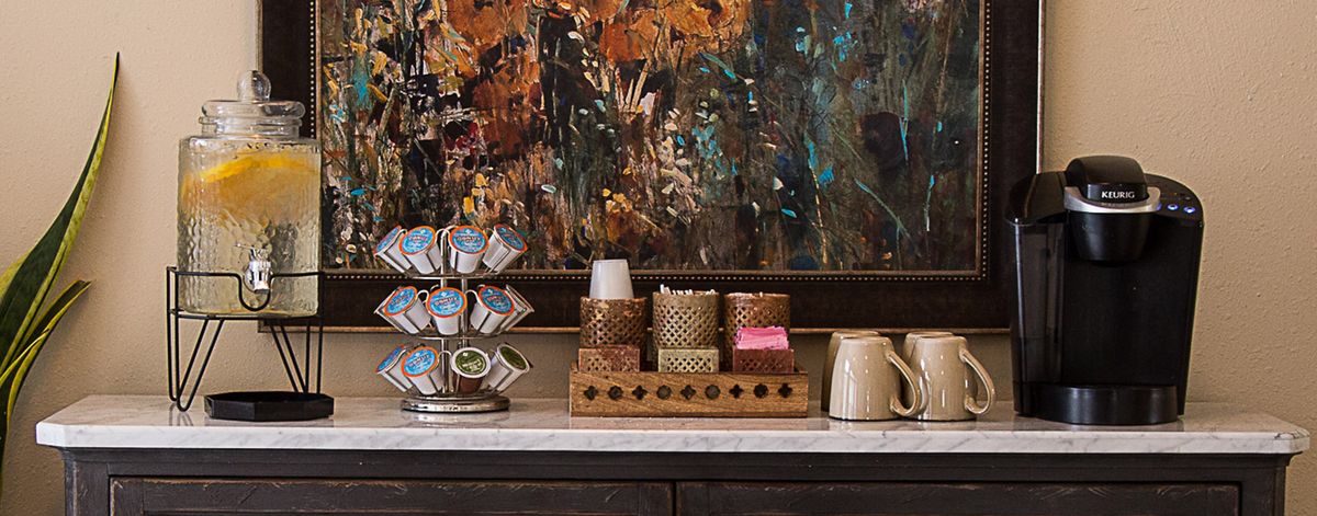 A cozy coffee station with assorted pods, mugs, and rustic decor against a colorful abstract painting.