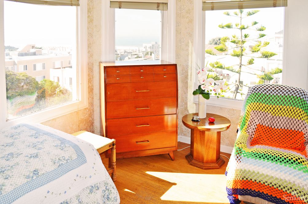 A cozy bedroom with natural light streaming through large windows, featuring a wooden dresser and round side table.