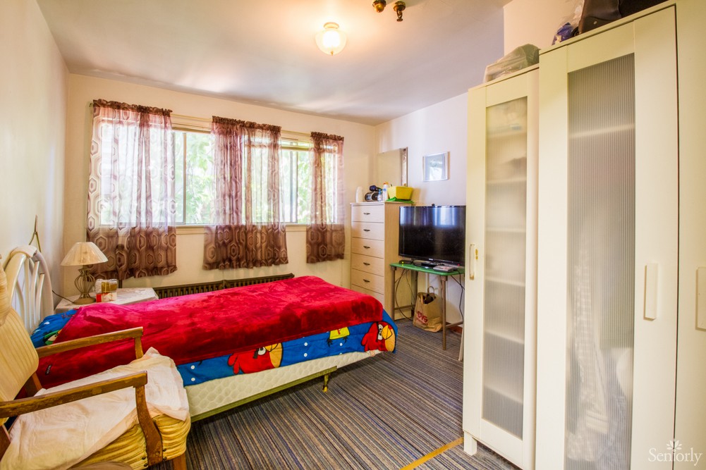 A cozy bedroom with a double bed and colorful bedding, featuring a television and wardrobe, warmly lit by natural light through large windows.