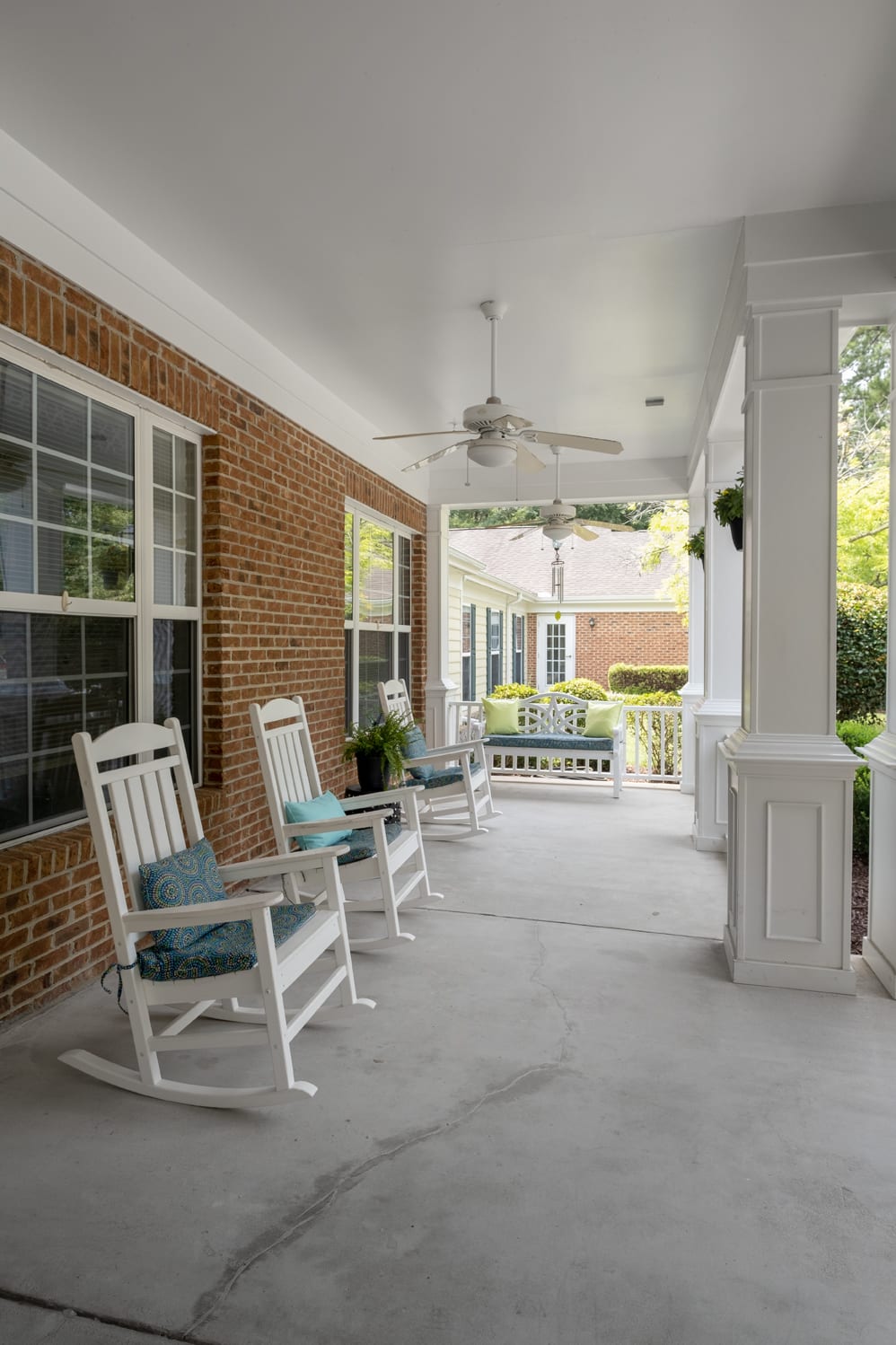 A cozy porch featuring white rocking chairs with cushions, a ceiling fan, and brick walls.