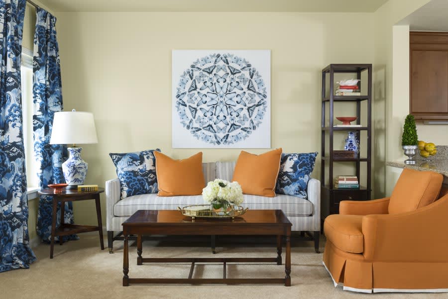 A stylish living room with a patterned artwork, comfortable sofa adorned with orange and blue cushions, a wooden coffee table, and an elegant shelving unit.