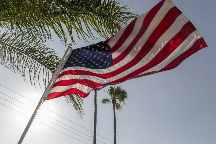 An American flag waves proudly under palm trees against a clear sky.