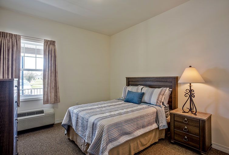A cozy bedroom with a double bed, nightstand, and lamp next to a window with curtains.