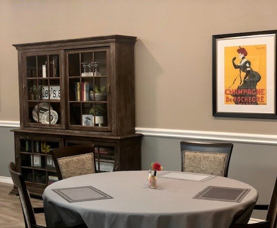 A cozy dining room with a round table, cushioned chairs, a wooden bookshelf, and wall artwork.