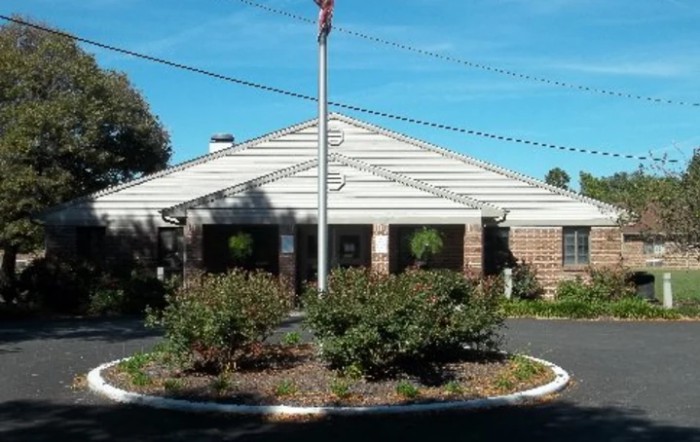 A building with a flagpole in front, surrounded by landscaping and trees under a clear sky.