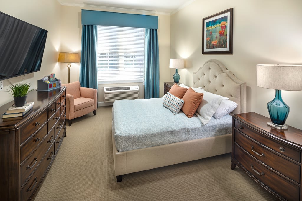 A cozy bedroom with a double bed, upholstered headboard, bedside tables, lamps, and an armchair near the window.