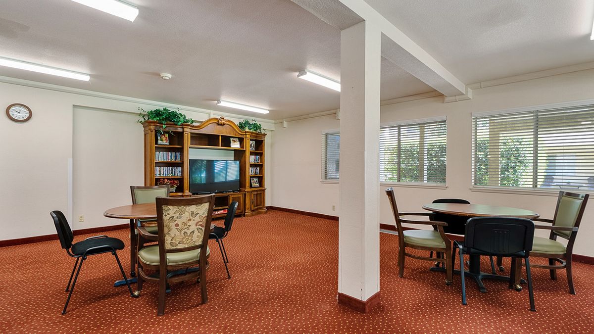 A cozy indoor room with a television, bookshelf, tables, and chairs on a carpeted floor.