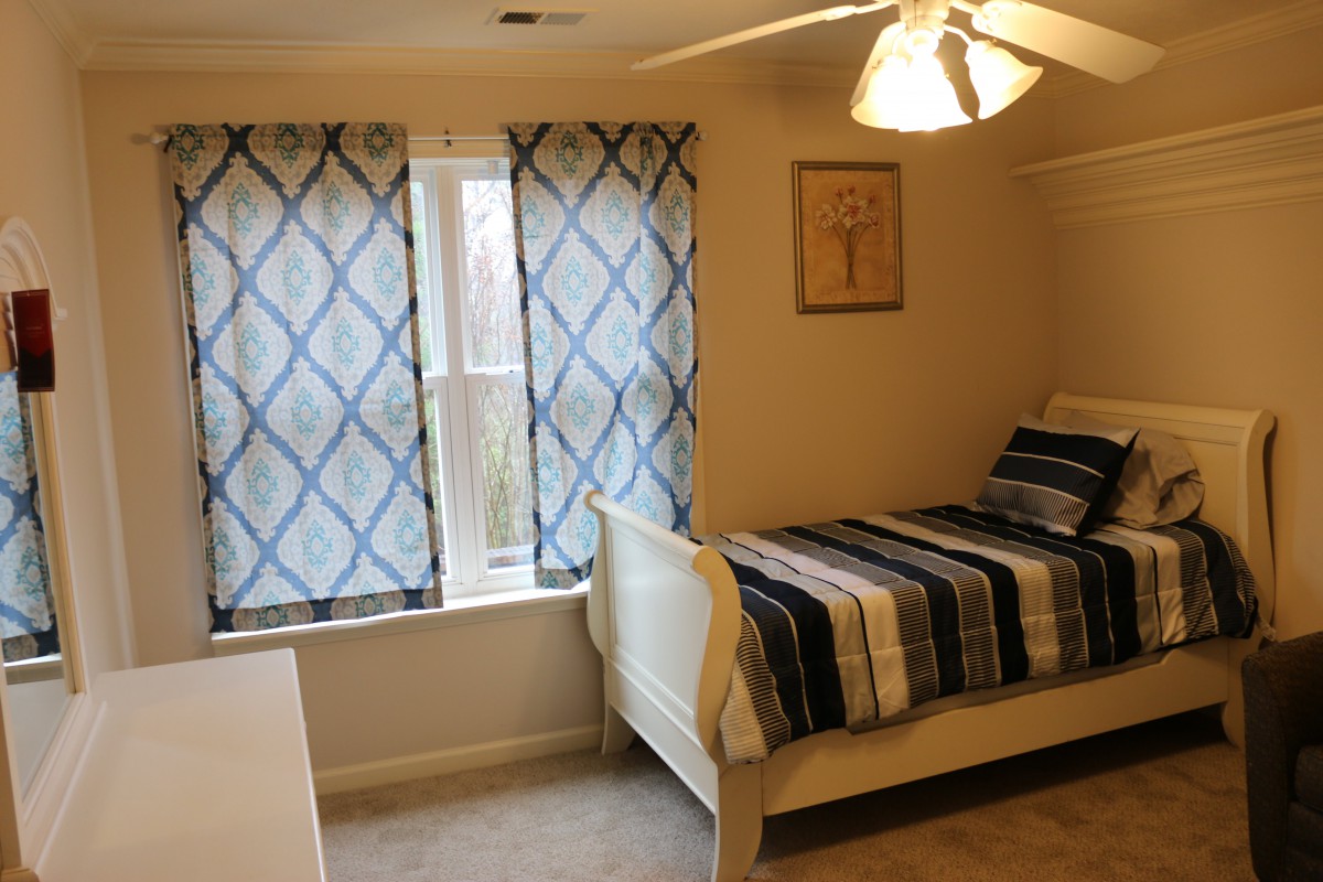 A cozy bedroom with a single bed featuring striped bedding, curtains on the window, and a ceiling fan.