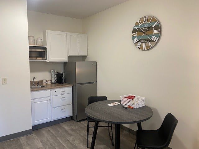 A kitchenette with a small dining area, featuring a table and chairs, fridge, microwave, cabinets, and a decorative wall clock.