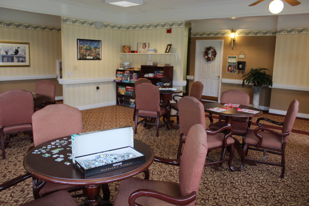 A cozy recreation room with tables, chairs, a puzzle in progress, and a bookshelf.