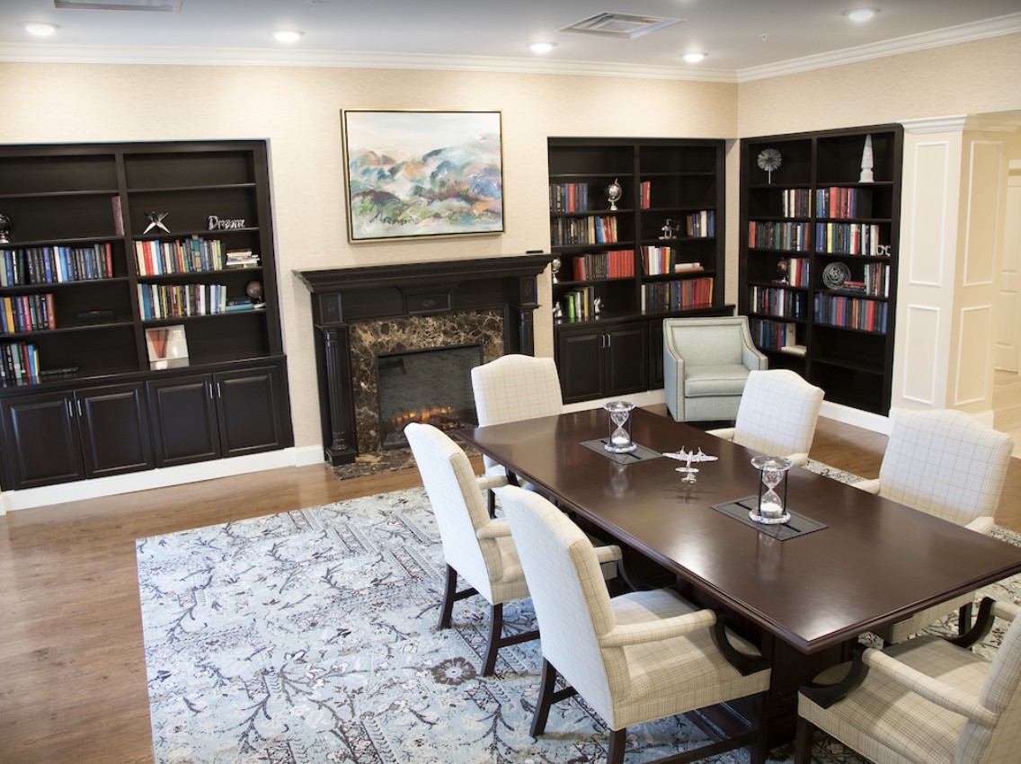 A cozy library room featuring a large wooden table and chairs, surrounded by bookshelves filled with books. A fireplace and artwork adorn the wall.
