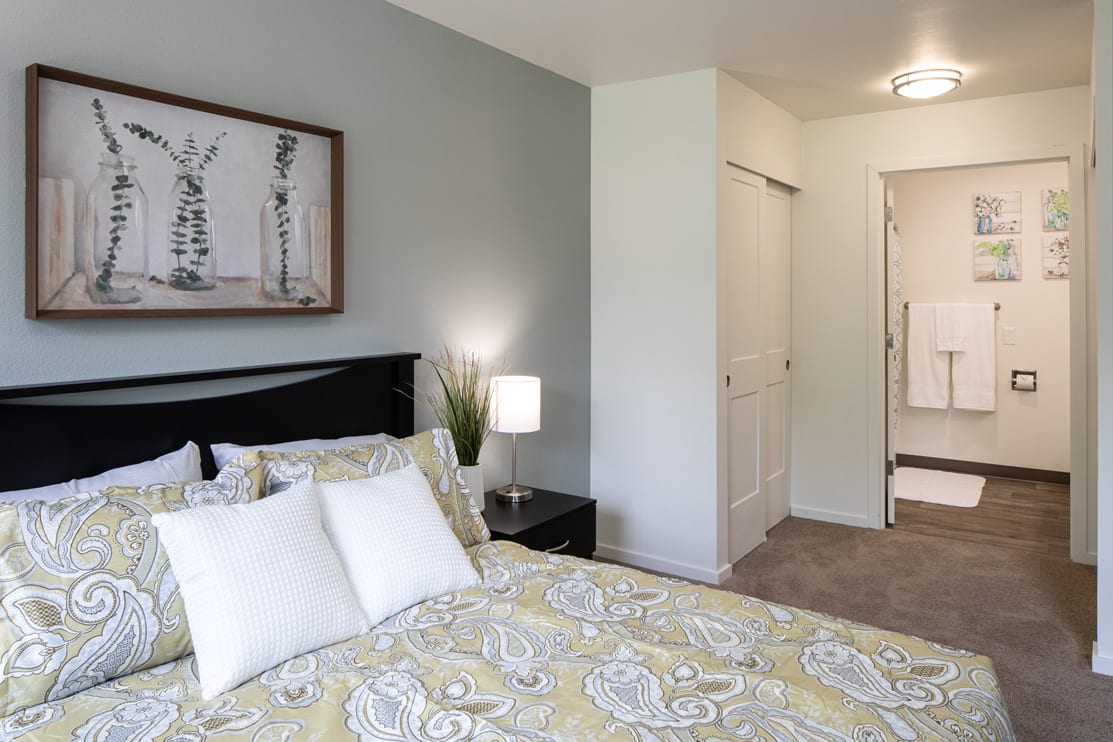 A modern bedroom with a neatly made bed, a nightstand with a lamp, and artwork on the wall.
