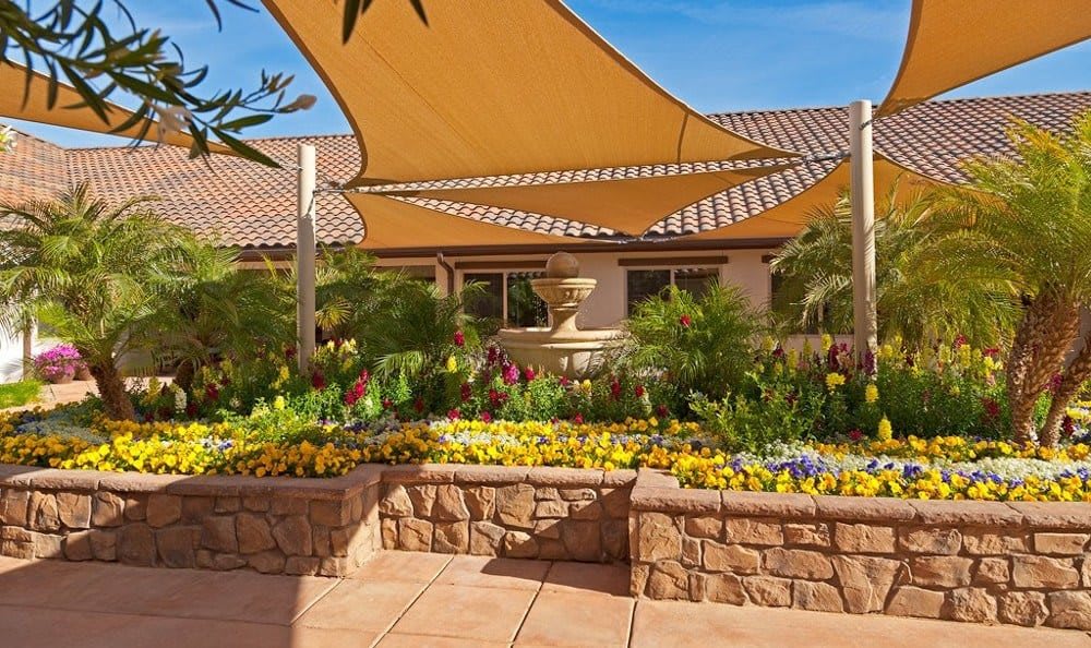 A tranquil courtyard features colorful flowers, a central fountain, and shade sails beneath a clear blue sky.