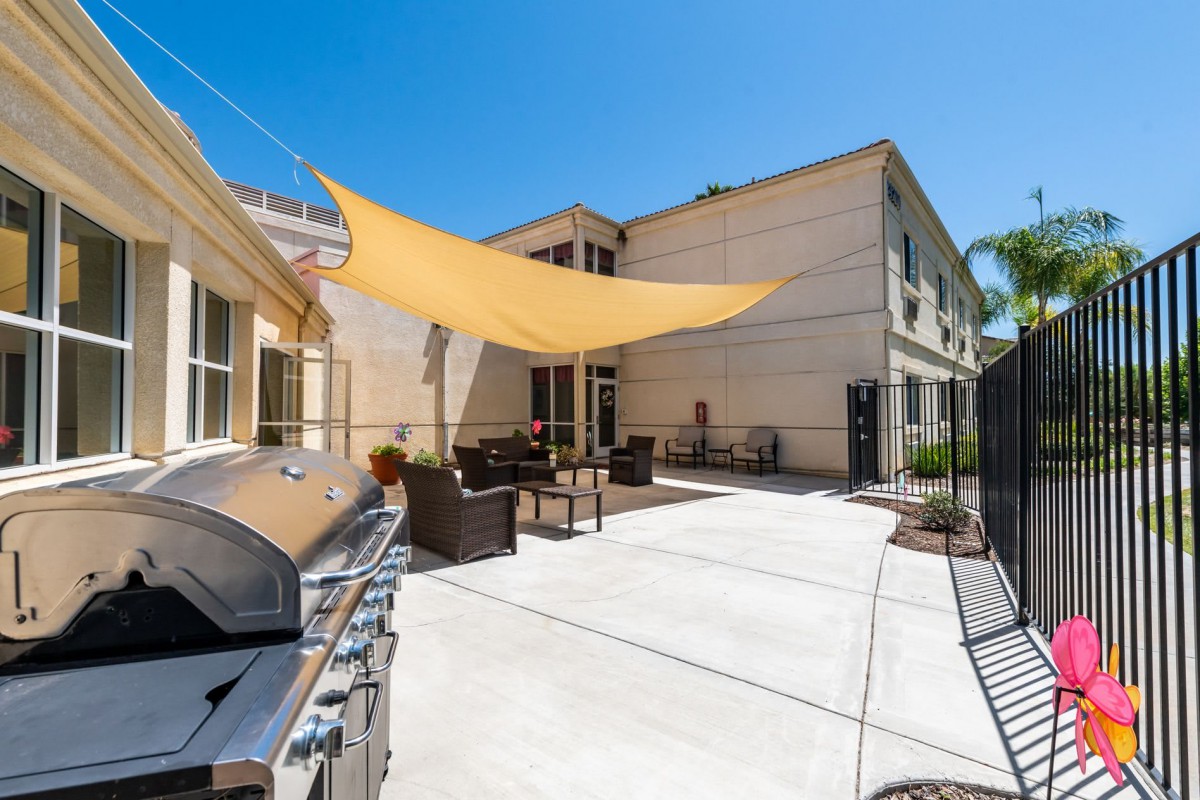 A sunny residential backyard with a patio, featuring a barbecue grill and outdoor furniture under a shade sail.