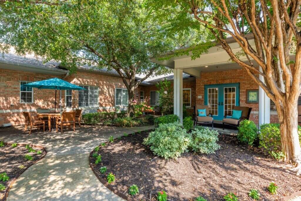 A serene garden patio with outdoor seating and shaded by trees, adjacent to a brick house.
