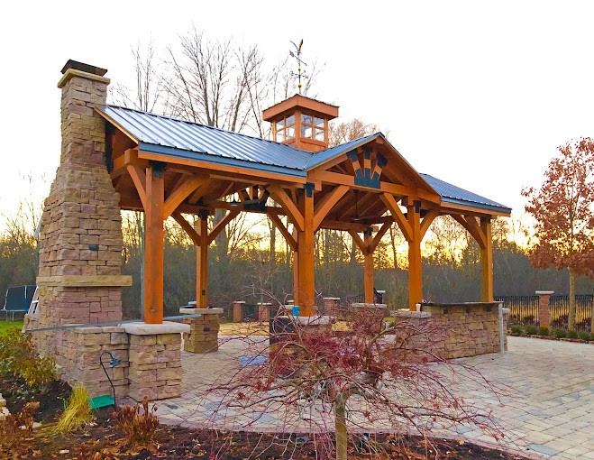 An outdoor timber frame pergola with a built-in fireplace, set against a natural landscape.