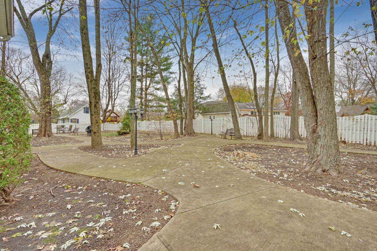 A spacious backyard with a concrete walkway surrounded by trees and a white fence.