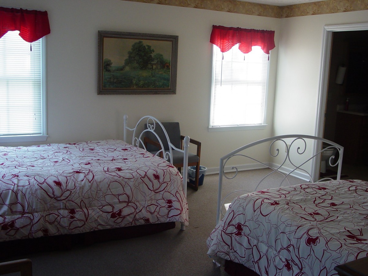 A cozy bedroom featuring two metal frame beds with floral bedding, a window with red valance, and a landscape painting on the wall.