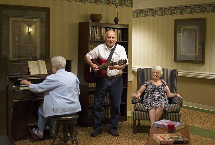 A man plays the guitar while a woman plays the piano and another woman sits in a chair, enjoying the music in a cozy living room setting.