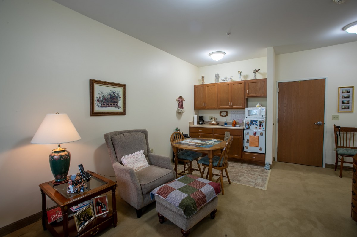 A cozy apartment living area with a kitchenette, featuring a comfortable chair, small dining table, and warm lighting.