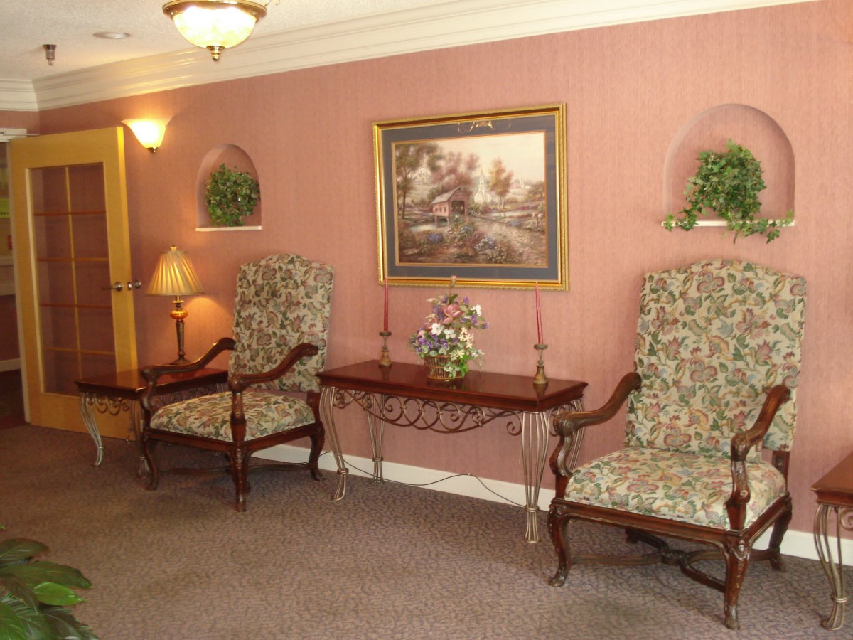 A traditionally styled interior with floral upholstered chairs, a wooden table, decorative plants, and a painting on the wall.