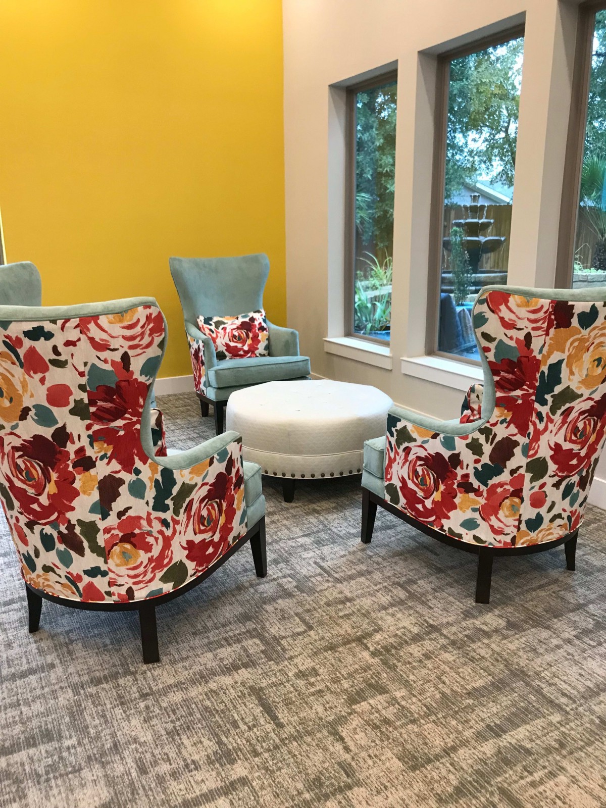 A cozy seating area with floral-patterned chairs and a round ottoman by large windows.