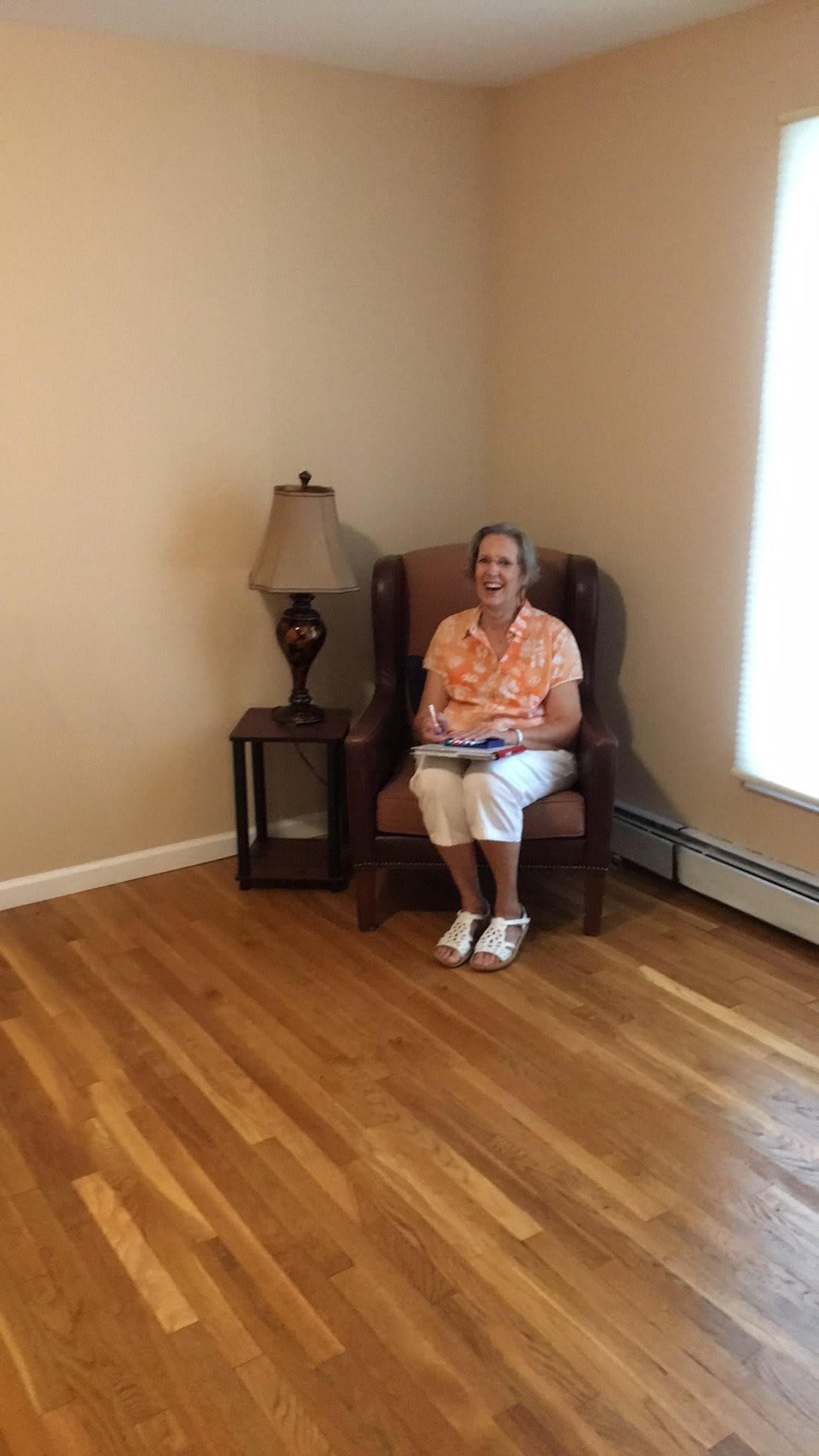 A woman sitting on a chair in a sparsely furnished room with wooden flooring.