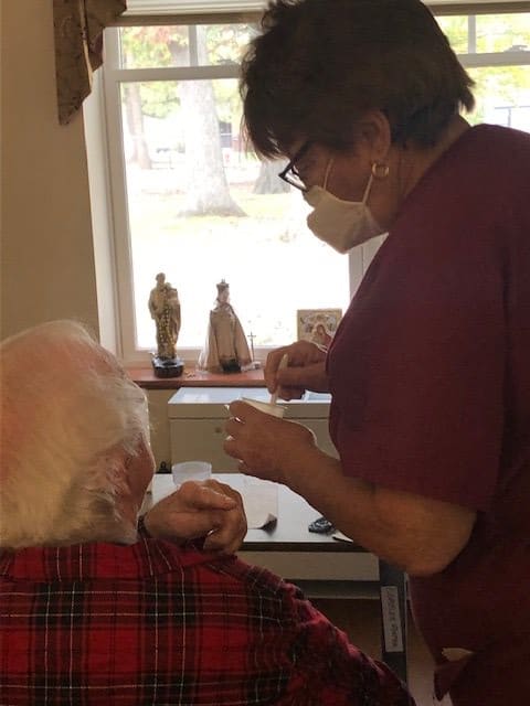 A healthcare worker providing assistance to an elderly person indoors, both wearing masks.