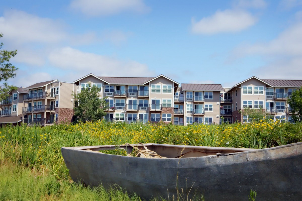 An abandoned boat rests in a grassy field with an apartment building in the background against a blue sky.