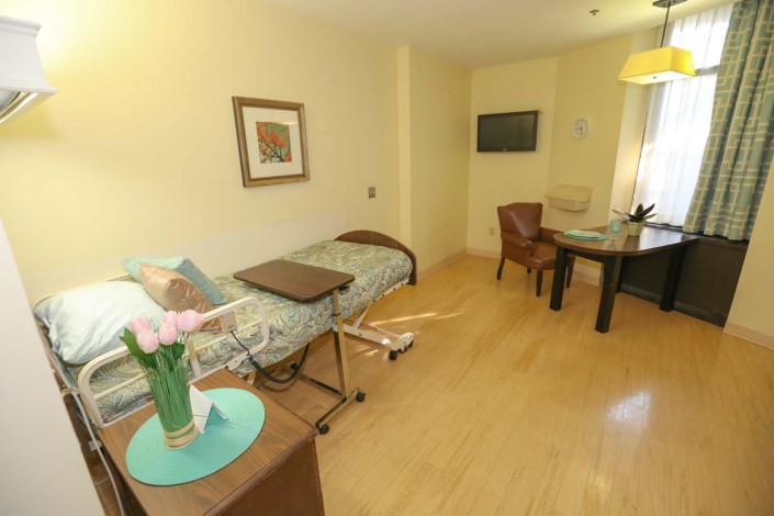 A well-furnished hospital room with a bed, table, chairs, and flowers on a side table.