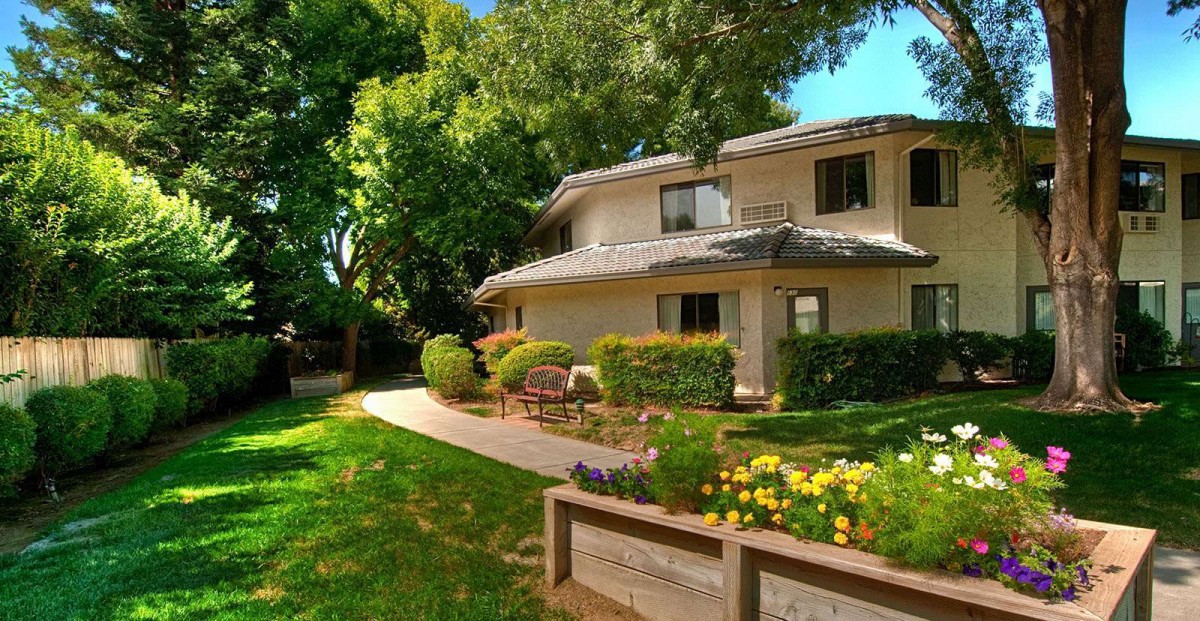 A two-story house surrounded by a lush garden with colorful flowers and tall trees on a sunny day.