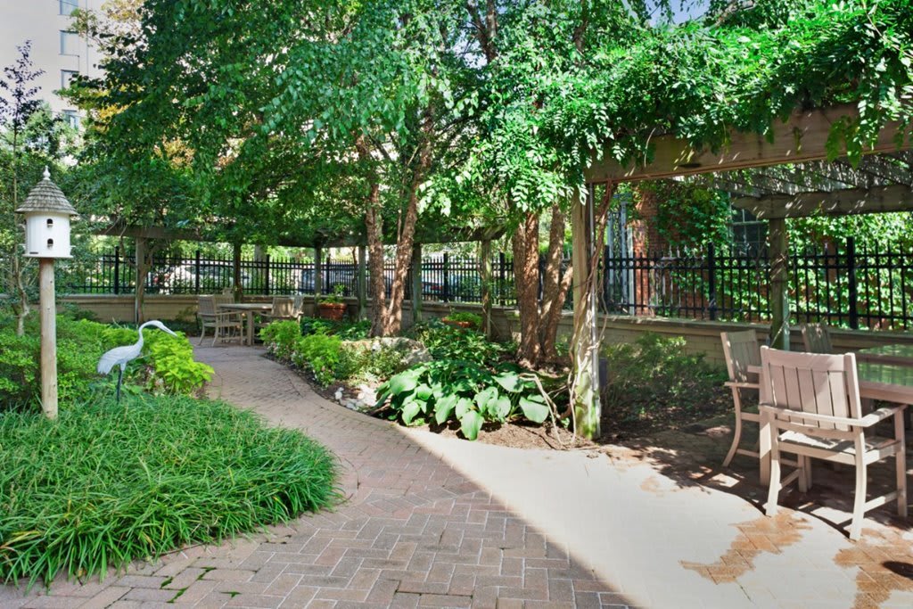 A peaceful garden pathway surrounded by lush greenery and shaded by trees, featuring a bench and a fence.