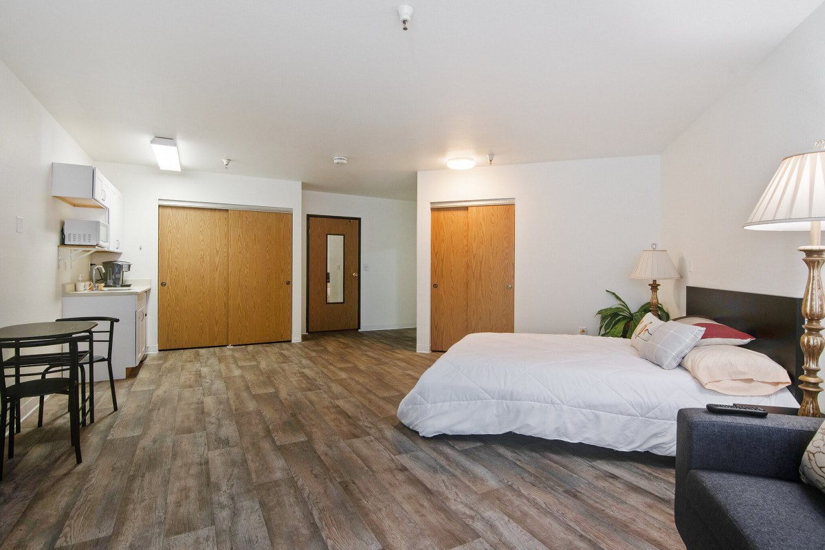 A minimalist bedroom featuring a double bed, wooden flooring, and modern lighting, complemented by an indoor plant and spacious wardrobes.