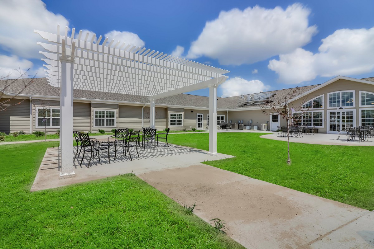A spacious outdoor patio area with a pergola and seating, surrounded by green lawn and under a clear blue sky with scattered clouds.