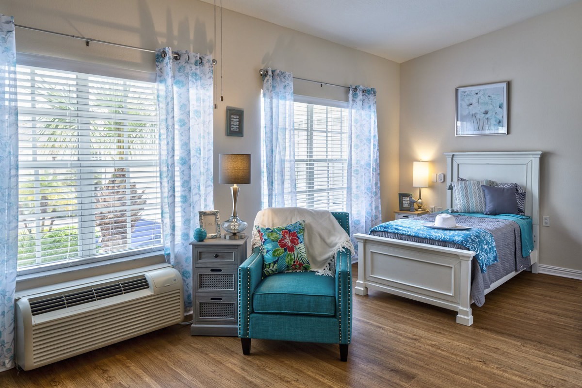 A cozy and well-lit bedroom featuring a blue armchair, elegant curtains, and wooden flooring.
