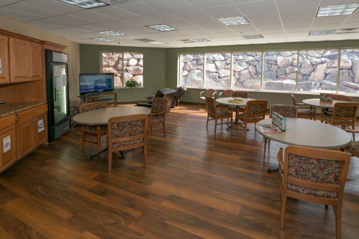 A spacious lounge area with wooden flooring, featuring several tables and chairs, large windows, and a television.
