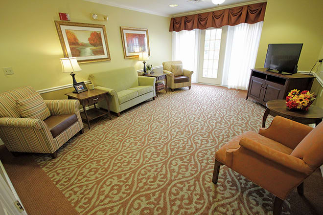 A cozy living room with sofas and armchairs arranged around a patterned carpet, adorned with paintings and ambient lighting.