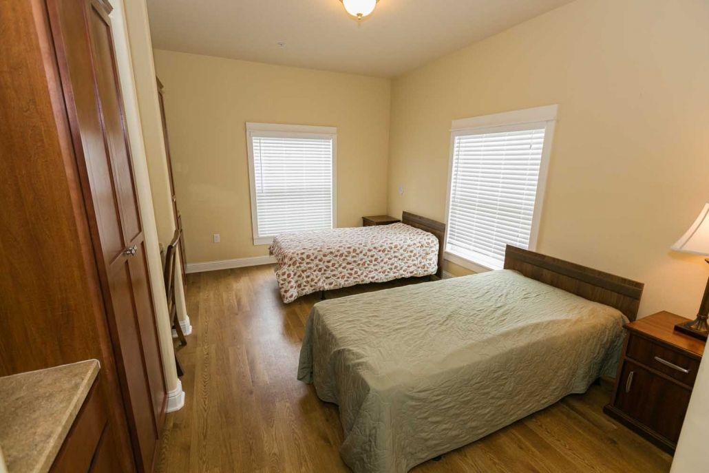 A simple bedroom with two beds and wooden flooring, featuring two windows for natural light.