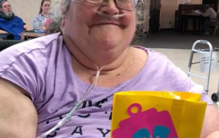 A smiling woman wearing a purple shirt and glasses, using an oxygen tube, holds a colorful bag.
