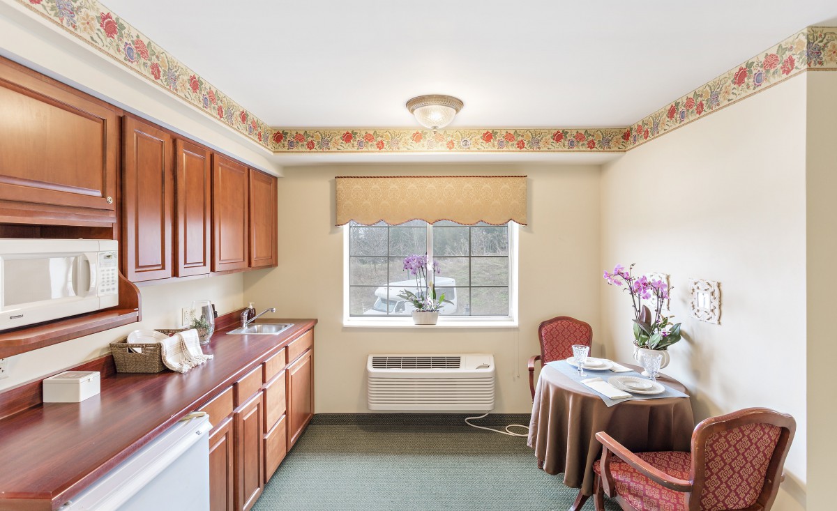 A cozy kitchen area with wooden cabinets, a small table and chairs, a window with flowers, and a neutral color palette.