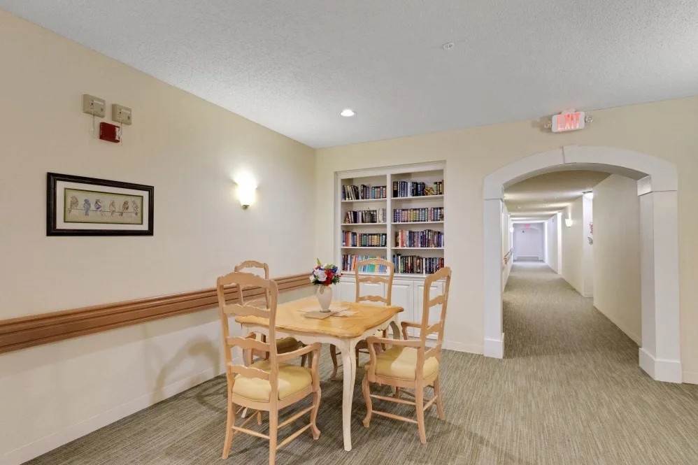 A well-lit room featuring a wooden table with chairs, a bookshelf, and an adjacent hallway.