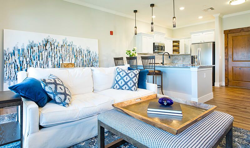 A modern living room with a white sofa, patterned cushions, and a view into an open kitchen area.