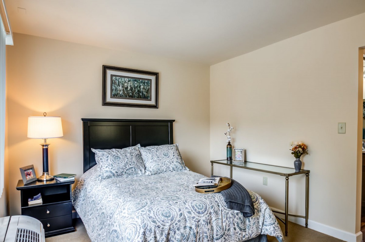 A cozy bedroom featuring a neatly made bed with patterned bedding, a nightstand with a lamp, and a desk against the wall.
