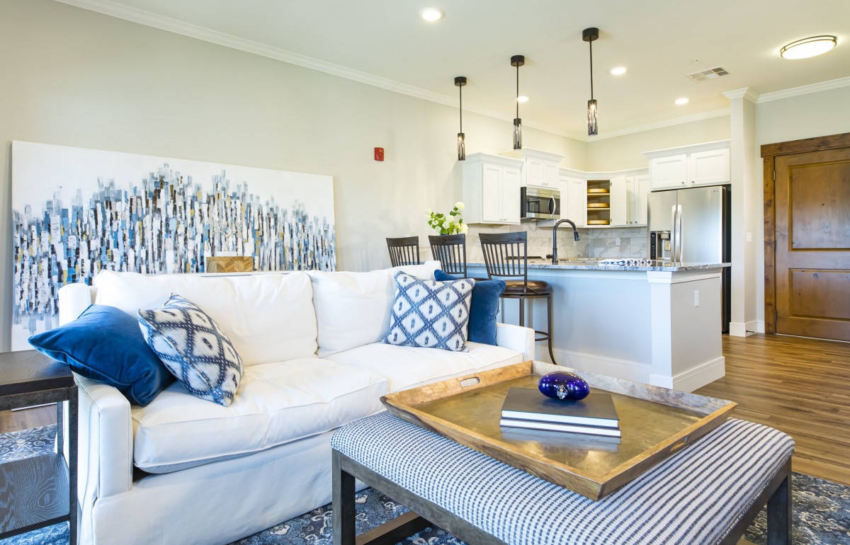 A modern open-concept living area featuring a stylish sofa with patterned cushions, a wooden coffee table, and an adjoining kitchen with bar stools and pendant lighting.