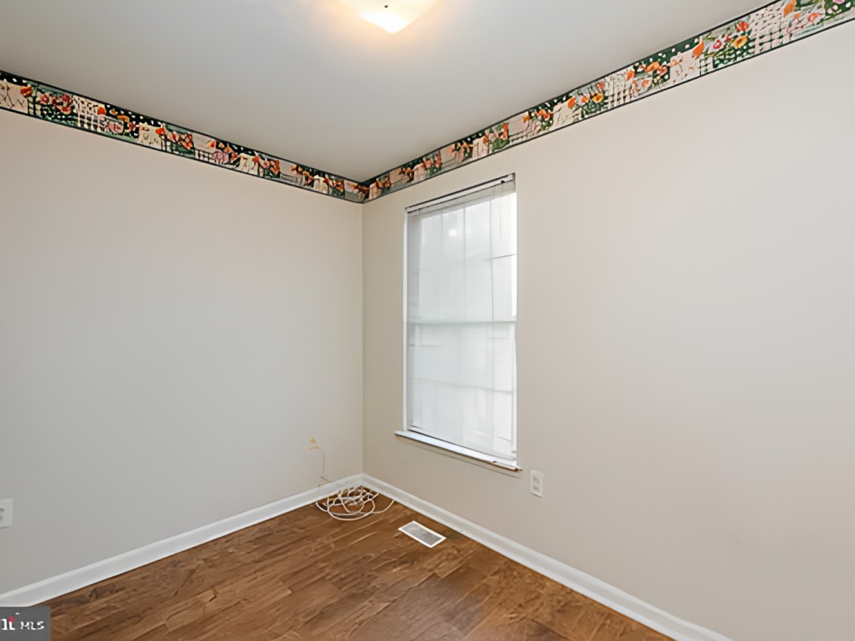 A small, empty room with a hardwood floor and a single window, featuring a wallpaper border near the ceiling.