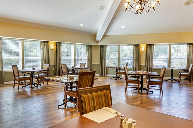 A spacious dining area with elegantly arranged tables and chairs, featuring large windows and a chandelier.
