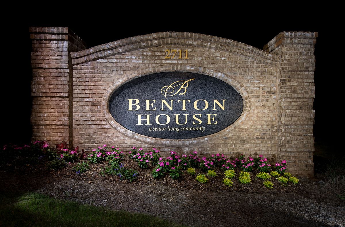 A well-lit Benton House sign is displayed on a brick wall surrounded by flowers at night.