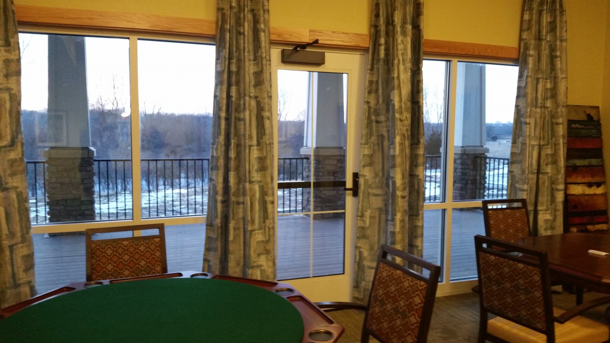 A room featuring a poker table and chairs, with curtains framing a sliding glass door leading to a balcony.