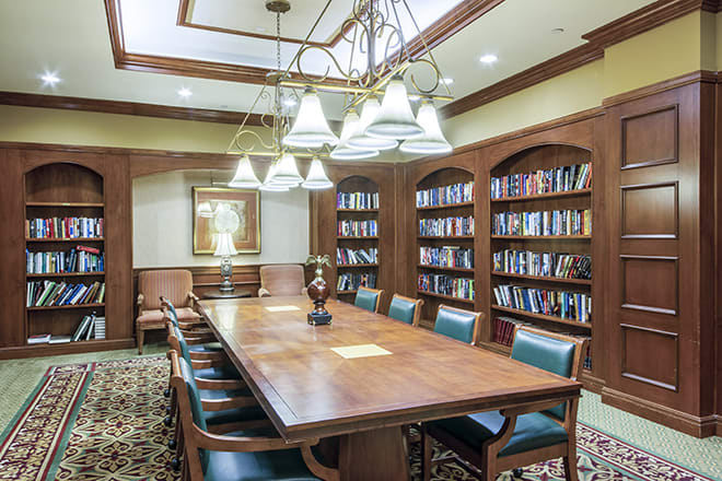 A well-lit conference room with a large wooden table surrounded by chairs, flanked by bookshelves filled with books.